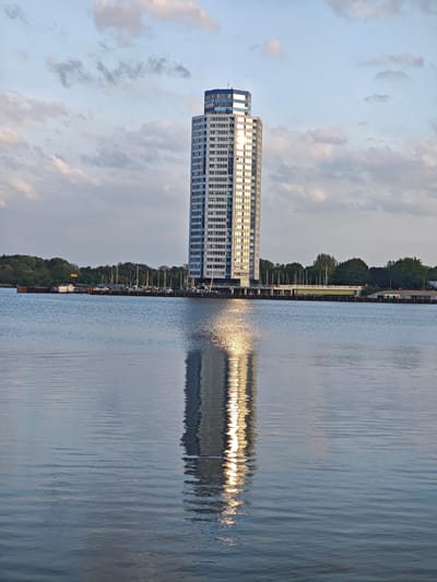 The image shows a tall, modern building, likely an office tower or hotel, situated on the edge of a body of water. The building is rectangular with a flat top, made of glass and concrete, and reflects the sunlight on its exterior. The water reflects the building, creating a mirrored image. In the background, there is a pier with docked boats and a line of trees. The sky is cloudy. The location is likely in Schleswig, Germany, as the building closely resembles the Holm-Passage in Schleswig.