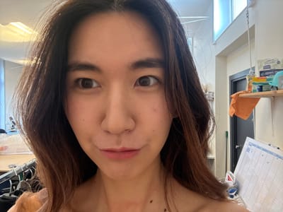 The image presents a close-up, selfie-style photograph of a young East Asian female in an indoor environment situated in Arcueil, France. The female subject has long, dark brown hair and light brown eyes, looking towards the right side of the frame with her lips slightly pursed. Moles are visible on her right neck and upper chest area. She is wearing an off-the-shoulder garment, appearing light peach or nude in color.
The background on the right features a light-colored wall with a partially visible dark door. A light wooden shelf is mounted on this wall, holding a yellow plastic bottle, a blue container, a smaller yellow container, and a light blue hard hat. An orange cloth is draped over the shelf's front edge. Below the shelf, a white grid board, marked with "X" in several cells, leans against the wall, next to a visible green mop or broom handle. Above the shelf and door, a high window or skylight is present, and a metal rod is suspended from the ceiling.
The background on the left is blurred but shows overhead fluorescent lighting fixtures, a clothing rack with light-colored garments on hangers, and a section of a workbench or table.