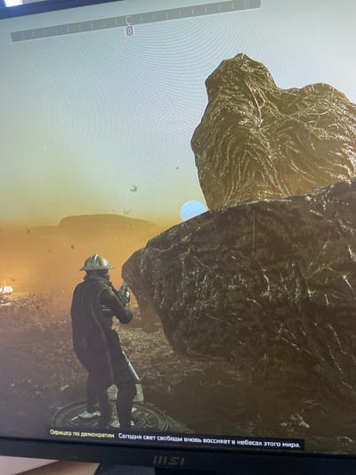 The image shows a video game scene with a person in a helmet and protective gear holding a weapon, standing on a platform in a rocky, alien-like environment. In the background, there are desolate landscapes with large rock formations and a hazy, orange sky. A small, pale blue orb is visible in the sky. Text in Russian appears at the bottom of the screen. The scene does not depict any location in the city of Beloretsk, Russia.