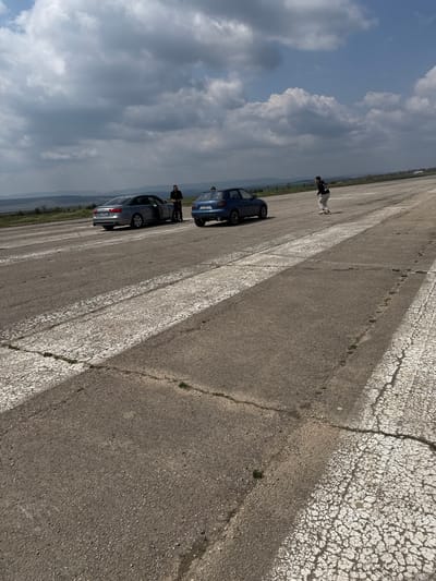 The image shows a cracked concrete surface, possibly a road or runway, in Buhovtsi, Bulgaria. Two cars, one blue and one silver, are parked on the left side. Two people stand near the silver car. A third person is standing at a distance away from the cars. The sky is cloudy.