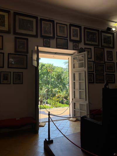The image depicts an interior view looking out through a partially opened white-framed window to a garden or outdoor space. The interior walls are adorned with numerous framed pictures of various sizes and styles, creating a gallery-like effect. On the right side of the frame, part of an object, possibly a piece of furniture or an exhibit stand with a red rope barrier, is visible. The window opens to a bright, green outdoor scene featuring lush vegetation, trees, and a distant railing. The natural light floods in through the window, contrasting with the darker interior. The location of the image is Bunyola, Spain.