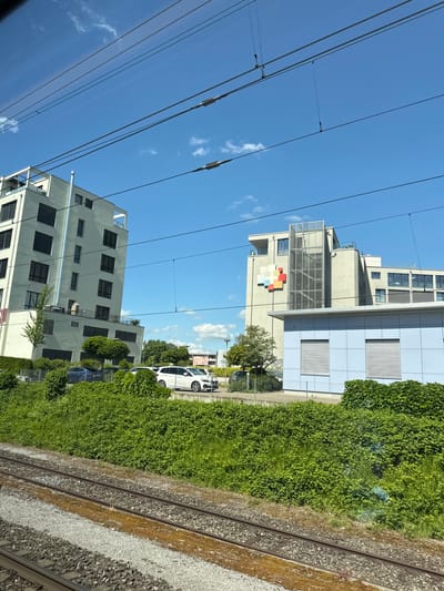 The image presents a detailed, forensic view of an urban landscape in Wetzikon (ZH), Switzerland, specifically facing the Spital Zürcher Oberland, observed from a railway perspective on a bright, sunny day.
In the lower foreground, two parallel railway tracks, laid on a bed of grey gravel ballast, extend horizontally across the frame, showing signs of light use with minor discoloration on the rails. Immediately beyond the tracks, a dense band of varied green vegetation, primarily thick bushes and taller grasses, forms a natural boundary between the rail infrastructure and the buildings.
The midground is dominated by modern multi-story buildings. On the right, a prominent light-grey building, identifiable as part of the Spital Zürcher Oberland, features a distinct multi-colored square logo on its facade, arranged in a cross-like pattern using blocks of red, orange, yellow, green, blue, purple, grey, and white. An external, glass-enclosed elevator shaft or staircase ascends the building's front. In front of this larger structure stands a smaller, light-blue rectangular building with two horizontal louvered windows. To the left of the hospital complex, another contemporary multi-story building, likely residential, presents a light-grey facade with numerous dark-framed windows and balconies, complemented by a tall, vertical silver pipe on its side. Between the vegetation and these buildings, a parking area is visible, accommodating several parked vehicles, including a white SUV and a blue sedan. Scattered trees and shrubs dot the landscape around the buildings and parking spaces.
The upper portion of the image reveals a clear, vibrant blue sky dotted with a few wispy white clouds. Multiple black and silver electrical overhead lines (catenary system) associated with the railway tracks crisscross the sky, suggesting an electrified rail line. No people are visible in the scene. The overall lighting is bright and clear, indicative of natural sunlight.