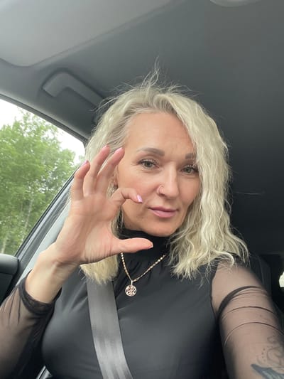 A woman with blonde hair is sitting in the driver's seat of a car, holding up her left hand in front of her face, with her fingers spread. She is wearing a black top with sheer sleeves and a gold necklace. The interior of the car is visible, including the dashboard and a gray seatbelt. Outside the car window, trees are visible, suggesting a rural or suburban setting. The woman appears to be looking at the camera. There is no specific indication of the location being Ul'yanovka, Russia.