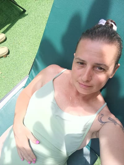 A woman is lying on a green chaise lounge outdoors on a sunny day. She is wearing a light green swimsuit and has her hair pulled back with a white hair tie. She has a tattoo on her left shoulder. The lounge is situated on a green artificial turf surface. A pair of sandals are visible on the turf. The location is Hua Hin City Municipality, Thailand.