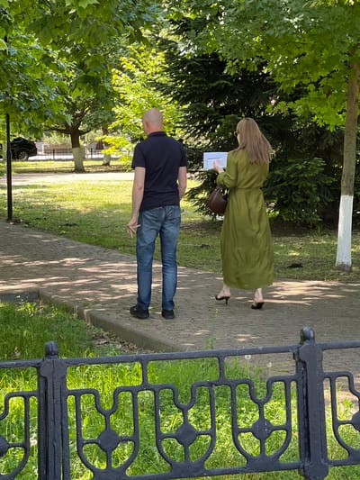 The image captures two individuals on a paved pathway in a park-like setting. A bald man, facing away from the camera, is wearing a dark blue polo shirt and blue jeans. He is standing on the right side of the pathway, with his left hand slightly raised. To his right, a woman with long, light brown hair, also facing away, is wearing a long, flowing olive-green dress and black heeled sandals. She is holding a white rectangular object, possibly a book or papers, in her hands. The pathway is bordered by green grass and a black metal fence in the foreground. Trees with lush green foliage and a dark green conifer tree are visible in the background. Sunlight filters through the leaves, creating dappled shadows on the path. The scene appears to be in Takhtamukai, Russia, indicated by the surrounding greenery and the style of the park infrastructure.