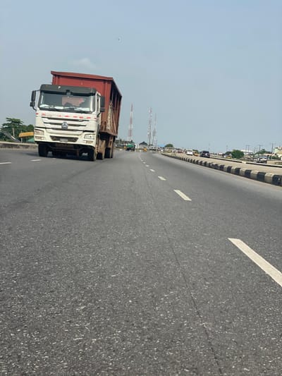 A wide, sunlit asphalt road with white lane markings stretches into the distance, occupied by a large white and red dump truck. Utility towers line the road further ahead, and a row of vehicles can be seen on a parallel road or shoulder to the side, potentially in a congested area. The sky is clear and bright, suggesting a sunny day.