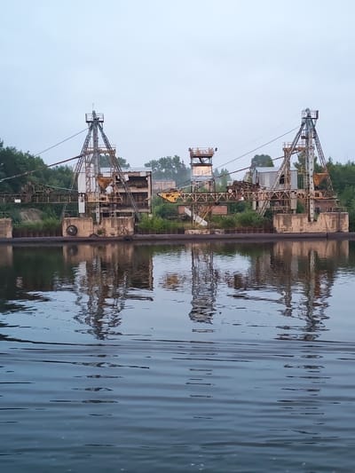 The image shows an industrial scene on a riverbank in Ivanov Bor, Russia. Two large, rusty excavators or cranes stand on a concrete structure along the water. The excavators are connected by cables and appear to be part of a dredging or mining operation. The water in the foreground reflects the structures, creating a mirror image. Trees and a small building can be seen in the background. The sky is overcast, suggesting a cloudy day. There are no people visible in the image.