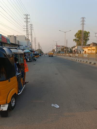 The image depicts a wide-angle street scene in Anantapur, India, captured during daylight under a clear, light sky. The asphalt road occupies the majority of the foreground and midground, appearing mostly clear of traffic with some debris, including a crumpled white object, on the surface.

On the left side, a prominent yellow and black auto-rickshaw is parked or stopped, facing away from the camera. Inside, a person wearing a blue shirt and dark flip-flops is seated. An orange fabric banner with black text in Telugu hangs from the auto-rickshaw's side. Behind it, a line of other auto-rickshaws, a green scooter, and a street vendor's cart with a light blue umbrella are visible. The left side of the road is flanked by a row of commercial buildings, some multi-story, displaying signage, including "SHEES" and text in Telugu script. Overhead, numerous power lines stretch across the sky, supported by multiple tall, metallic electricity transmission towers that run parallel to the road.

The right side of the road features a central median with a black and white curb, bordered by a yellow metal barrier. Beyond the barrier, further down the street, are various buildings, including a light yellow structure and a multi-story building with the partial word "HOSPITAL" visible. A large, leafy green tree stands beyond these structures.

In the distance, a yellow mini-truck or utility vehicle is visible on the right side of the road, heading away from the camera. Scattered individuals are visible walking or standing on both sides of the road in the far distance. Streetlights with multiple arms are positioned along the median and sides of the street. The overall environment appears urban with a mix of commercial and potentially residential structures.