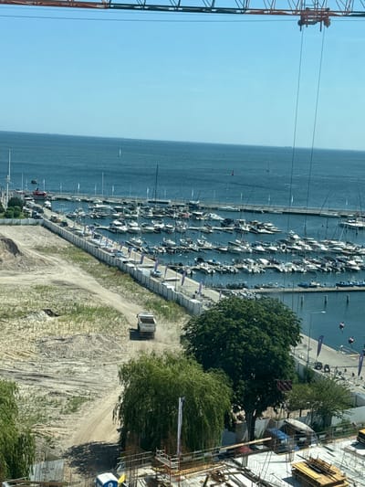 This image shows a construction site adjacent to a marina in Gdynia, Poland. In the foreground, a construction site is visible with scaffolding, building materials, and a portable toilet. A dirt road leads from the construction site towards a marina. A white dump truck is driving on the dirt road, kicking up dust. The marina is filled with numerous sailboats and motorboats docked in the water. Beyond the marina, the Baltic Sea stretches out under a clear blue sky. The horizon shows a distant coastline with what appear to be ships. A large crane is visible in the upper portion of the image, indicating ongoing construction work.