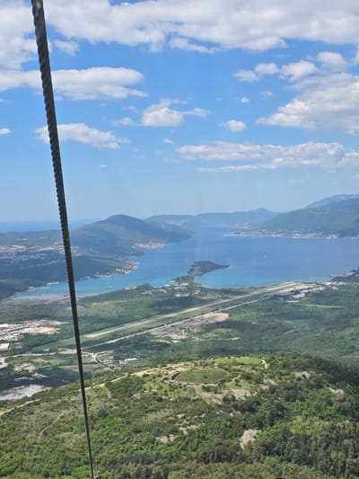 A slightly tilted wide outdoor shot from a high vantage point depicts Mirac, Montenegro, on a sunny day with scattered clouds in a blue sky.

The bottom-left corner of the image features a twisted steel cable. To the right of the cable, the view extends to a landscape divided into three main areas: a body of water, a shoreline with built infrastructure, and a tree-covered mountainous region. The water stretches far into the distance and is dotted with boats, with a small island visible in the middle. The shoreline, adjacent to the water, features a long road and stretches along the edge of the tree-covered mountain. Buildings are sparsely distributed along the road and shoreline. The mountainous region is densely covered with green trees.