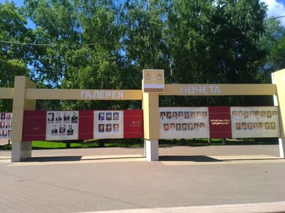 The image shows a monument with portraits of people displayed on panels. The monument has a yellow superstructure with the words "ГАЛЕРЕЯ ПОЧЕТА" (Gallery of Honor) written on it in white letters. Below this, there are red panels with portraits and text in Russian, such as "ГЕРОИ ТРУДА" (Heroes of Labor) and "ПАТРИОТЫ ОТЕЧЕСТВА" (Patriots of the Fatherland). Further to the right, there are more panels with portraits and the text "ПРЕДСТАВИТЕЛИ ПРЕДПРИЯТИЙ" (Representatives of Enterprises). The monument is set against a backdrop of lush green trees and a blue sky. The ground in front of the monument is paved with light-colored bricks. The monument is located in the city of Otradny, Russia.