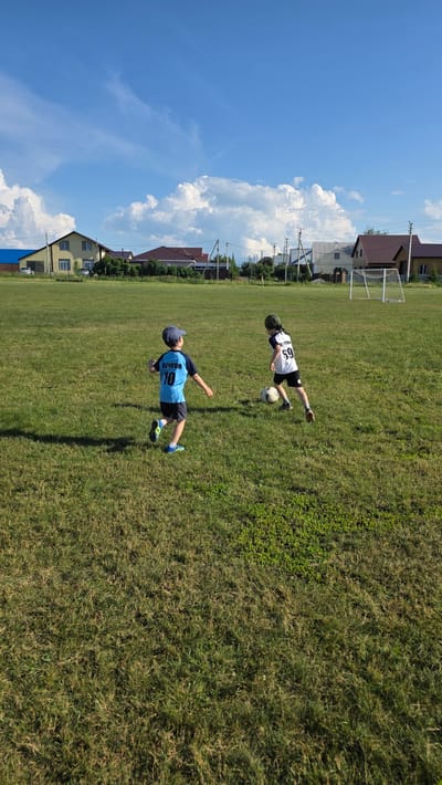 In Bulgakovo, Russia, two young boys are playing soccer on a grassy field under a bright blue sky with scattered white clouds. The boy on the left wears a blue t-shirt with the number 10 and the name "БУЧКОВ" on the back, dark shorts, a blue cap, and blue cleats. He is in motion, running with his arms outstretched. The boy on the right is dressed in a white t-shirt with the number 59 and dark shorts, and he is wearing a dark cap. He is actively engaged with the soccer ball at his feet. In the background, several houses with pitched roofs and a soccer goal are visible. The field is well-maintained grass with some patches of longer grass. The overall scene captures a casual outdoor activity on a sunny day.