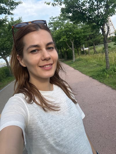 A woman with brown hair and sunglasses perched on her head smiles into the camera while standing on a paved pathway in a park. She is wearing a white t-shirt. The pathway is lined with green trees and grass. In the background, a modern white structure is visible behind the trees, and a body of water can be seen in the distance to the left. The scene appears to be in Gimpo-si, South Korea, given the park setting and architectural style.
