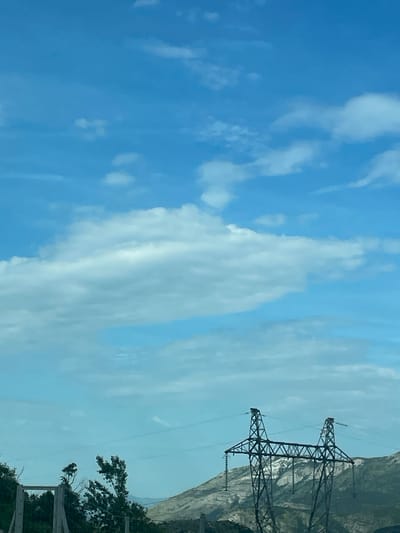 The image presents a vertically oriented view of a partially cloudy, blue sky. In the upper right, two electrical transmission towers are visible against a light-colored, rocky hillside. In the lower right corner, a tree, green foliage, and parts of a fence are present. It is impossible to say if the location is in the city of Bolesestra, Montenegro.