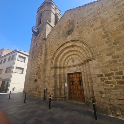 The image shows a stone church in La Jonquera, Spain, viewed from a low angle. The church has a bell tower with a clock on its side. The building is made of tan-colored stone blocks. The front facade features a large arched doorway with a wooden door and a round window above it. A three-story building with windows is visible to the left of the church tower. The foreground shows a paved street with black bollards. The sky is blue.
