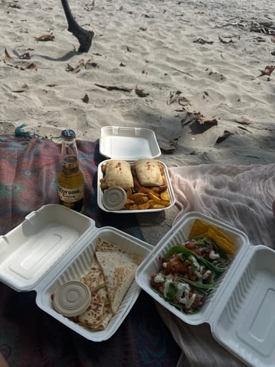 The image shows a picnic on a beach in Santa Fe, Costa Rica. The scene features a picnic blanket laid out on the sand. On the blanket are three take-out containers, each holding different food items. One container has two sandwiches with potato wedges, another has a quesadilla, and the third has three tacos. There is a bottle of Corona beer and a brown paper cup lid in the photo. The containers are white with a paper-like texture. The beach is sandy with a few scattered leaves and the shadow of a tree branch can be seen in the upper left portion of the image. The blanket appears to be a colorful fabric with pink, blue, purple, and green tones. The picnic is set up on the beach, facing the ocean. The beach is located in Santa Fe, Costa Rica, with coordinates 9.642 latitude and -85.168 longitude.