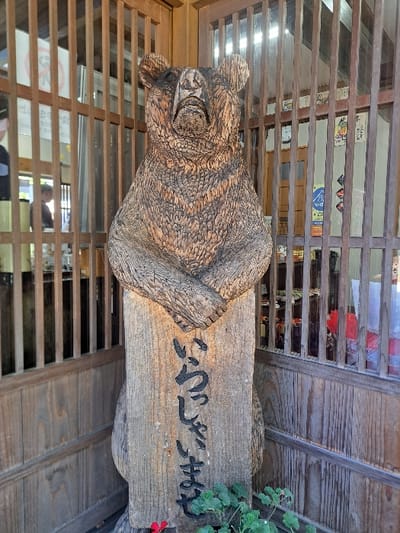 A light brown wooden bear carving stands upright in a corner of a wooden-paneled structure, likely an entrance or interior of a commercial establishment in Shirakawa, Japan. The bear, carved with textured fur details, has its forepaws crossed over its chest and its head slightly tilted upwards. Its lower body transitions into a rectangular wooden base bearing the vertically carved Japanese inscription "いらっしゃいませ" (Irasshaimase), meaning "Welcome." The bear is situated in front of vertical wooden slats forming a screen or partition. Through these slats on the left, a blurred figure is partially visible within an interior room, alongside a white sign. On the right, more interior space is visible through the slats, showing colorful signs and items on shelves. A small potted plant with green leaves and one red flower is positioned at the base of the bear. The scene is evenly lit, casting soft shadows and highlighting the wood grain.