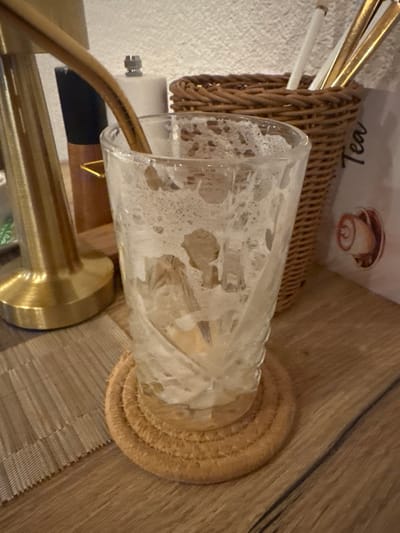 The scene depicts an indoor setting, likely a cafe or restaurant in Visp, Switzerland. In the foreground, an almost empty clear glass, featuring a textured or fluted exterior, rests on a circular, light brown braided coaster made of natural fiber. The glass contains residual light brown liquid and white foam, consistent with a recently consumed beverage such as a milkshake or iced coffee. A thin, gold-toned metallic straw or stirring spoon extends into the glass from the upper left.

To the left of the glass, a prominent golden, reflective metallic object, possibly the base of a lamp or a decorative stand, is visible on a grey striped placemat. Behind this object, a portion of a black and brown condiment grinder is discernible. To the right of the glass, a brown woven wicker basket holds several gold-toned metallic utensils or straws, alongside a white plastic straw. Behind the basket, a white menu or sign is partially visible, displaying "Tea" vertically and a circular graphic resembling a cinnamon roll. All objects are positioned on a light brown wooden table exhibiting a visible grain. The background wall is light-colored and textured. No individuals are present in the image.