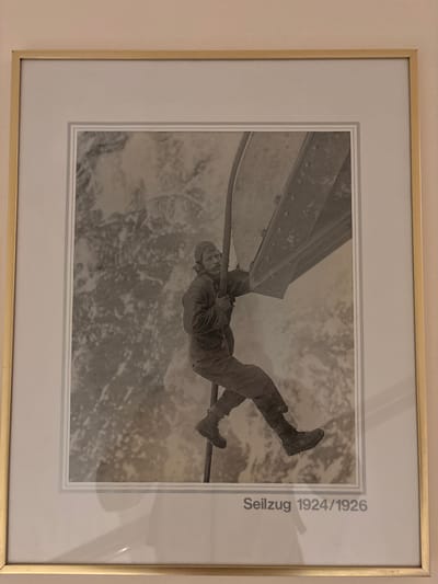 The image is a framed black and white photograph showing a man climbing on a thick cable. He is wearing outdoor clothing including a hat and heavy boots. He is holding onto the cable with his hands and has his legs wrapped around it. In the background, there is a blurry, mountainous landscape covered in snow. The photograph is titled "Seilzug 1924/1926" which translates to "Cable Car 1924/1926". This title, combined with the snowy mountainous terrain, suggests the photograph was taken in a high-altitude location, possibly in connection with the construction or operation of a cable car system. Given that the title refers to a period between 1924 and 1926 and mentions "Seilzug" which can also refer to a cable railway, it is plausible that this depicts an early stage of a cable transport system in Ehrwald, Austria, as Ehrwald is known for its mountainous terrain and cable car facilities.