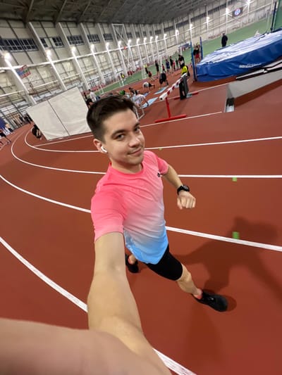 A man in athletic wear takes a selfie on an indoor running track. He wears a pink and blue gradient shirt, black shorts, and running shoes. A watch is visible on his left wrist, and he has a wireless earbud in his ear. The track is red with white lines, and hurdles are set up on the left side. Other athletes are visible in the background. The building has a high ceiling with many lights. The image is slightly tilted.