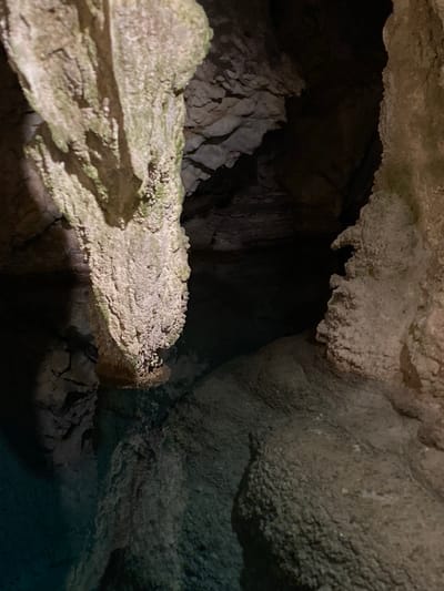 The image depicts a dark, subterranean cave environment. The foreground is dominated by rough, textured rock formations, some appearing as stalactites or stalagmites. A prominent, light-colored formation with a rough surface hangs from above, casting shadows. To the left, a body of dark, possibly water, is visible, reflecting the dim light of the cave. The background recedes into deep shadow, suggesting a large, unexplored cavern. The textures of the rock are varied, ranging from smooth and worn to jagged and crystalline. Greenish moss or algae is visible on some of the rock surfaces, indicating moisture. The overall impression is one of a natural, ancient, and somewhat mysterious setting. No people, objects, or text are visible. The image conveys a sense of depth and the raw beauty of geological formations. The lighting is dim and atmospheric, typical of an underground cave.