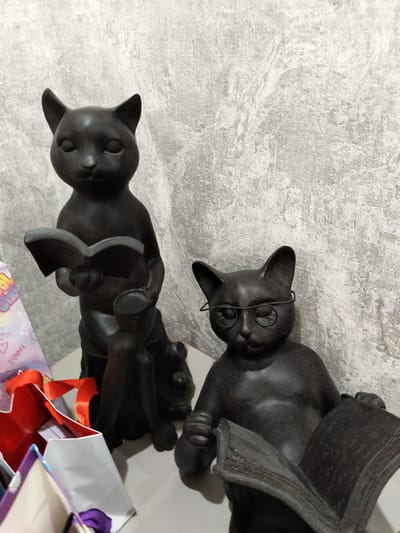 Two black cat figurines are displayed on a light-colored surface in front of a textured gray wall. The figurine on the left is sitting and holding an open book. The figurine on the right is also sitting, wearing round wire-rimmed glasses, and holding an open book. Various gift bags, some with red ribbon, are visible in the foreground. The scene appears to be indoors, with artificial lighting. There is no clear indication of time of day or weather. One of the gift bags has the word "FUNNY" printed on it in pink lettering. The overall tone is decorative and whimsical, with the cat figurines depicting a scholarly and studious theme.