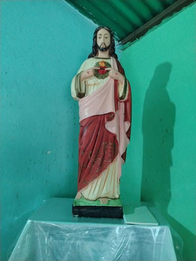 A full-length statue of Jesus Christ stands on a table covered with a clear plastic tablecloth in the corner of a room with light-green walls. Jesus is depicted with long, dark hair and a beard, wearing a cream-colored robe draped with a pink and red cloth. His hands are raised, pointing towards a stylized heart with flames emanating from it on his chest. A notebook lies on the table next to the statue's base. A tall, dark shadow of the statue is cast on the right wall. There is no indication of the statue being in Los Leoncitos, Venezuela.