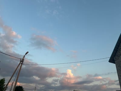 The image captures a twilight scene with a pale blue sky dotted with wispy clouds tinged with pink and gray. In the foreground, a tall, weathered utility pole stands at an angle, supporting electrical wires and a streetlamp. The pole is made of concrete and has a visible support structure. To the right, the corner of a building constructed of light-colored bricks and topped with a dark sloped roof is visible. The overall impression is of a residential or semi-rural area in Нижние Муллы, Russia, during the early evening. No people are present in the frame.