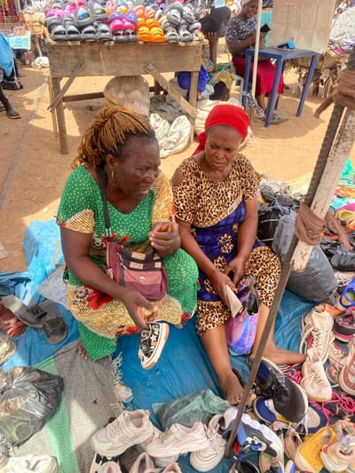 This image captures an outdoor market scene in Makera, Nigeria, under bright daylight.

In the foreground, two adult women are seated barefoot on a blue tarpaulin spread on the dirt ground, surrounded by various types of footwear for sale. The woman on the left wears a green and yellow patterned dress with cut-out shoulders and has light brown braided hair. A maroon shoulder bag is slung across her body. She holds a small, black-and-white patterned canvas shoe in her right hand, her head tilted downwards as if examining it or speaking. The woman on the right wears a red headscarf and a leopard-print top with a blue patterned wrapper. She holds a light-colored item, possibly cash or paper, along with a dark object resembling sunglasses within an iridescent purple bag on her lap. Numerous new and used shoes, including sneakers, sandals, and slippers of various colors and designs, are displayed around them on the tarpaulin. A black plastic bag rests on the left side of the tarpaulin.

In the midground, a rustic wooden table displays rows of colorful rubber sandals and slippers. Behind this table, another individual, partially obscured, is visible. Further into the background, a person wearing a white headscarf is seated at a small blue table, focused on a handheld device. The overall environment features a sandy or dirt ground, indicative of an open-air market, with other patrons and simple stall structures visible in the distance.