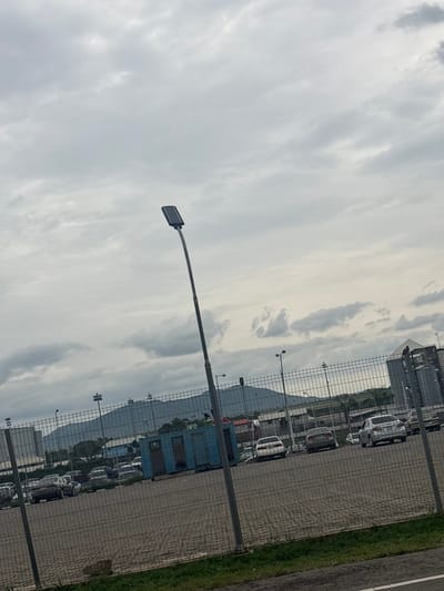 The image depicts an outdoor scene with a sky full of clouds. In the foreground, a tall lamppost with a modern, rectangular light fixture is visible, extending diagonally across the frame. Beyond the lamppost, a chain-link fence runs horizontally, separating the viewer from the rest of the scene.
Through the fence, a parking lot filled with numerous white cars is observable. The cars appear to be new or in pristine condition, possibly indicating a car dealership or storage facility. In the background, there are several buildings, including what looks like a blue container or modular building, and a larger structure with windows. Mountains or hills are faintly visible in the distance, shrouded in a light haze.
The weather appears overcast or cloudy, suggesting it might be morning or late afternoon, or simply a cloudy day. The lighting is diffuse and muted. The setting seems to be a commercial or industrial area, possibly a car storage yard or a manufacturing plant, located near a more natural landscape with hills. There is a strip of green grass and an asphalt road or pathway to the right of the fence. No people are visible in the image, and no text is discernable. The overall impression is one of a functional, somewhat utilitarian, outdoor space under a cloudy sky.