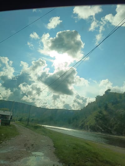 This image depicts a natural landscape within the city of Gabdyukovo, Russia. The scene is illuminated by daylight under a partly cloudy sky, with the sun partially obscured by a central cloud formation, creating distinct sunbeams and glare. The sky is light blue, populated by numerous white and grey cumulus clouds. Multiple dark utility cables stretch across the upper sky.
The foreground features a muddy dirt road with visible tire tracks and several puddles, one exhibiting a clear sky reflection. Green grass lines the road's edges. On the far left, a partial view of a white vehicle's bumper and a wooden structure, possibly a house, are discernible, along with two wooden utility poles supporting overhead wires.
A river or large stream flows diagonally from the mid-right, displaying surface reflections of sunlight. The riverbank is covered in green vegetation. Beyond the river, steep, heavily forested hills and mountains dominate the background, with a prominent solitary tree visible on the summit of the rightmost hill. No human or animal subjects are present. The environment combines natural elements with subtle indications of rural infrastructure.
