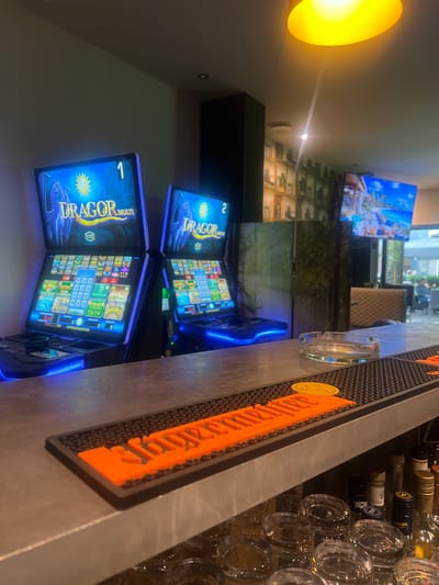 The image depicts the interior of a bar or casino in Ludwigshafen am Rhein, Germany. Two "DRAGOR MULTI" slot machines are prominently featured, illuminated by blue neon lights. The machines display various game interfaces. In the foreground, a bar counter is visible, with an ashtray and a "Jägermeister" branded bar mat. Behind the bar, shelves stocked with bottles and glassware can be seen. A television screen displaying a tropical scene is mounted on the wall in the background. The lighting is dim, with a bright yellow light fixture visible in the upper part of the frame. The environment appears to be a commercial establishment, likely catering to gaming and entertainment.