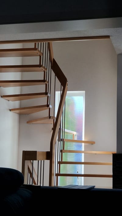 This image shows an interior view of a modern wooden staircase with a unique, slatted design. The staircase features light-colored wooden uprights and railings. The steps are not visible, but the structure suggests a split-level design or a landing area. The staircase is set against a white wall. A long, horizontal window runs along the upper part of the wall, offering a view of greenery outside. The window is covered with a textured, frosted glass that diffuses the light, creating a colorful, almost pixelated effect with hues of blue, green, and pink. The light coming through the window suggests it is daytime, possibly late afternoon or evening due to the warm lighting.

The scene appears to be in a residential setting, characterized by its clean lines and minimalist aesthetic. The overall mood is calm and contemporary. The image is taken from a slightly elevated viewpoint, looking down and across the staircase. The foreground shows a dark silhouette, possibly a piece of furniture or an architectural element, on the right side of the frame, partially obscuring the view of the lower part of the staircase. The design of the staircase, with its open slatted structure, allows light to pass through, contributing to a bright and airy feel. The location is identified as Ichenhausen, Germany, which might suggest a European architectural style. There is no visible text in the image.
