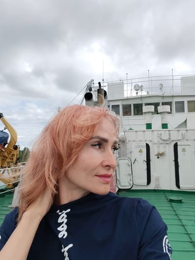 The image shows a woman with pink hair standing on the green deck of a ship. She is wearing a dark blue t-shirt with white writing on it. The ship appears to be docked or moving slowly, with a large white structure behind her that includes windows, doors, and ventilation systems. The sky is overcast. A crane-like structure is visible on the left side of the frame. The location is identified as Vytegra, Russia.
