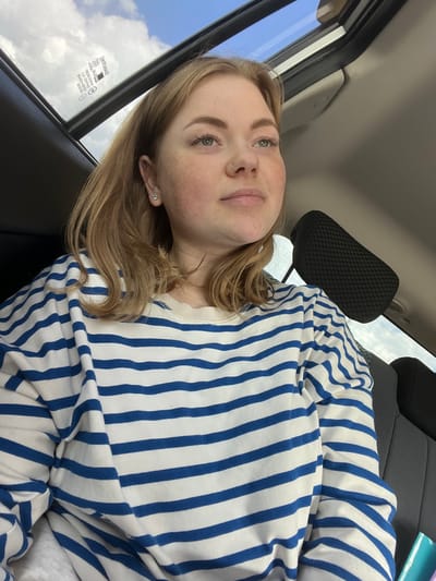 A young woman with blonde hair and light-colored eyes is pictured in a selfie taken inside a vehicle. She is wearing a blue and white striped long-sleeved shirt. She has a nose ring and earrings. The background shows the interior of a car, including a headrest and part of a seat. Through the window on the left, a cloudy blue sky is visible, suggesting it is daytime. Some text is visible on the car window, but it is not legible. The image is oriented sideways.