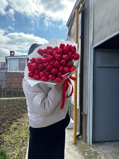 A person is standing outdoors in Trudobelikovskiy, Russia, holding a large bouquet of red tulips that completely obscures their face and upper torso. The individual is wearing a light beige puffer jacket and dark pants. The bouquet, consisting of numerous red tulips, is wrapped in white paper and clear plastic, secured with a prominent red ribbon. To the left of the person, a patch of tilled earth with nascent green shoots is visible, backed by a brick building featuring a window and a chimney. To the right, a textured building wall is visible, with a vertical yellow utility pipe running downwards and a dark gray roller blind partially covering an entrance. A gray metal utility box is affixed to the lower section of this wall. The ground beneath the person consists of a concrete or tiled surface, bordered by sparse grass. The sky above is blue with scattered white clouds.