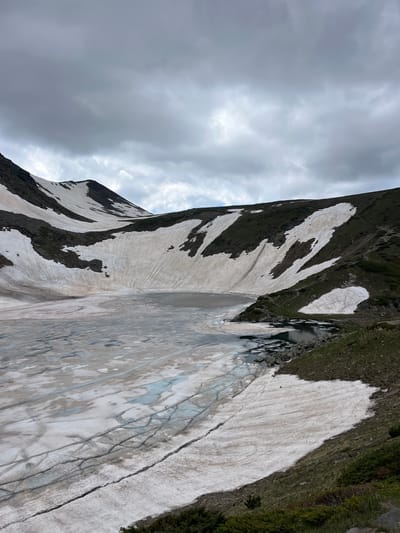 The image depicts a mountainous scene in Panichishte, Bulgaria, with a dramatic overcast sky dominating the left side. The terrain is rugged, with dark, rocky slopes interspersed with patches of snow. The snow is not uniformly white but shows streaks of dirt and meltwater, suggesting a thawing period. In the mid-ground, a body of water, possibly a lake or pond, is visible, with rocks scattered along its edge. The right side of the image shows a steep, grassy incline with sparse vegetation. The overall impression is one of a wild, untamed landscape under a cloudy sky. There are no people or man-made objects visible. The time of day is difficult to ascertain due to the overcast conditions, but it appears to be daylight. The weather is cool and potentially damp, with heavy clouds indicating a possibility of rain. The location is clearly mountainous, with the snow patches suggesting a higher altitude. No text is visible in the image.