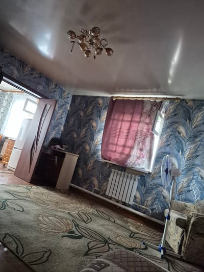 This is an indoor photograph taken in the city of Sokur, Russia, depicting a room. The room has patterned wallpaper with a floral or leaf motif in shades of blue, grey, and white. The ceiling is white and slightly reflective, with a modern chandelier featuring multiple glass globes. Natural light streams in from a window, casting reflections on the ceiling.
In the foreground, a large rug with a floral pattern in muted greens, beiges, and browns covers the floor. To the left, a tall, brown wooden door is leaning against a dresser that appears to be white and dark brown. Adjacent to the dresser is a white refrigerator. A window is visible behind the door and refrigerator, with light curtains, one a deep pink, and another with a floral print. A white radiator is situated below the window.
To the right of the frame, a white standing fan with blue accents is positioned near a patterned armchair. The armchair has a beige and brown floral upholstery. The overall impression is of a lived-in domestic space.