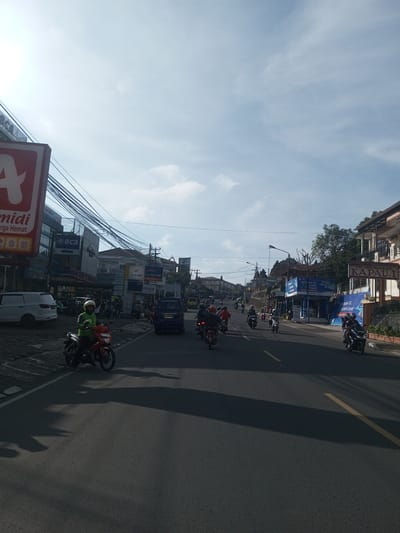 The image captures a street scene with a high volume of motorcycle traffic on a sunny day. Buildings line both sides of the road, displaying a mix of commercial and residential structures. Storefronts are visible, with signs indicating businesses such as banks (BCA, bank bjb), a pharmacy (mia farma), a clinic, and a restaurant (RUMAH MAKAN KAPAU.I). A large advertisement for "TRUST CELL" is also prominent. Overhead, power lines are dense and sprawling. The location appears to be in a bustling urban area. There is no indication of being in the city of Phou Khoun, Laos.