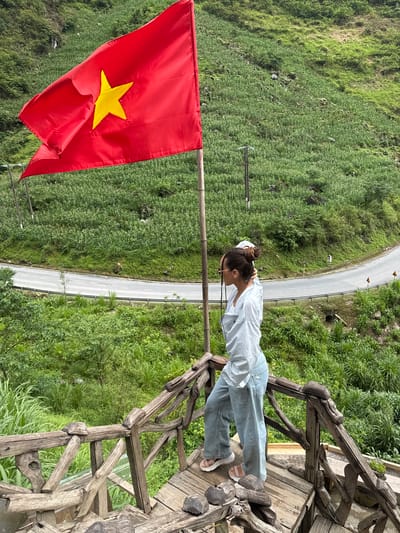 A woman is standing on a rustic wooden structure next to a road in Yên Minh Commune, Vietnam. She has her hair up, is wearing sunglasses, a light-colored shirt, and jeans. The woman is looking toward a large Vietnamese flag that is waving in the wind. The flag is red with a yellow star. The background consists of a steep, green hillside and a road winding through the landscape.