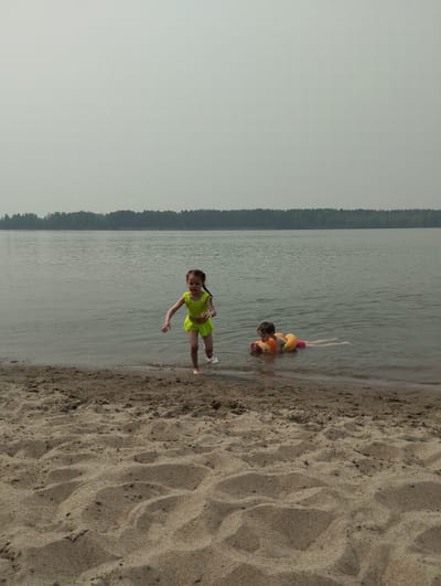 In a wide shot of a serene lake on a hazy, possibly overcast day, two young children are enjoying the water. The scene is set on a sandy beach, with the water's edge lapping at the shore. A dense line of trees forms a dark horizon across the far side of the lake.
On the right side of the image, a child is floating on their back in the water, wearing an orange and yellow inflatable swim ring around their torso and what appears to be a pink swim bottom. They are looking upwards.
To the left, a child in a bright yellow swimsuit, consisting of a ruffled top and a skirted bottom, is running out of the water onto the sand. Their arms are outstretched, and they appear to be in mid-stride, splashing as they exit the water.
The overall mood is one of innocent childhood play and summer fun, despite the muted lighting suggesting it's not a bright sunny day. The location appears to be a natural body of water with a sandy embankment, typical of a recreational area. There is no visible text in the image.