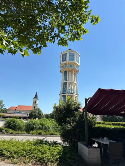 The image captures a low-angle view of a tall, multi-tiered observation tower in Siófok, Hungary, framed against a clear blue sky. The tower has a pale yellow structure with multiple glass-enclosed viewing platforms. Green foliage from a tree occupies the lower left corner of the frame. Adjacent to the tower, partially obscured by greenery and an umbrella, are tables and chairs that suggest a cafe or outdoor dining area. In the background, a church steeple rises above a building with a red roof, adding to the urban context. The scene is brightly lit, indicating a sunny day. A paved pathway bordered by grass and low shrubs runs along the right side of the image, leading the eye towards the background elements.