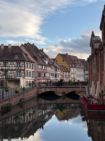 The image depicts a picturesque canal in the Petite Venise district of Colmar, France. The canal is flanked by colorful half-timbered buildings with steeply pitched roofs. The architecture suggests a German influence. A stone bridge crosses the canal, and people are visible walking across it. On the right side of the canal, a red-colored structure with umbrellas and tables suggests an outdoor seating area for a restaurant or cafe. The water reflects the buildings and the sky, creating a mirror-like effect. The sky above is partly cloudy with streaks of white clouds against a blue background. The overall scene evokes a charming and tranquil atmosphere typical of European canal towns.