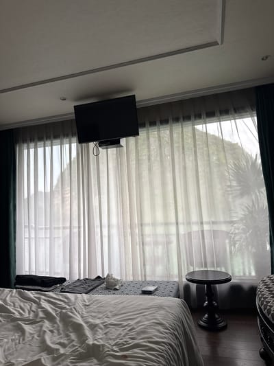 The media file shows an indoor scene, likely a bedroom, in Hải Phòng, Vietnam. The room is decorated with a mix of modern and possibly traditional elements, suggested by the detailed molding on the wall and the ornate pedestal table. A large window is covered by sheer white blinds, through which the outlines of trees, possibly palm trees, are visible, suggesting it is daytime and the weather might be overcast or the blinds are diffusing the light.
Dominating the left side of the frame is a white wall with decorative molding. Mounted on this wall is a flat-screen television. To the right of the television, the sheer blinds are the most prominent feature, covering a large window.
In the mid-ground, to the right, a black, pedestal-style side table with a round top is visible. Next to it, a bed with rumpled white linens takes up the right side of the image. A small, patterned headboard or bed frame is partially visible between the bed and the blinds. Some dark clothing is draped over the side of the bed or a nearby surface.
The overall impression is one of a hotel room or a well-appointed bedroom. The lighting is soft and diffused, typical of an indoor setting with natural light coming through blinds. There is no visible text in the image.