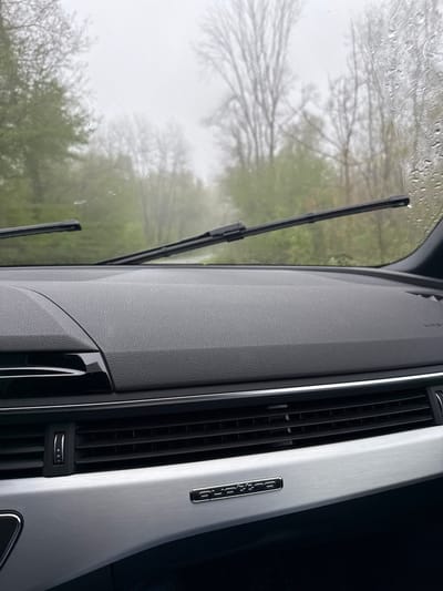 The image shows the interior of a car from the driver's perspective. The car is driving on a road surrounded by trees. It is raining, as evidenced by the raindrops on the windshield and the windshield wipers in motion. The dashboard of the car is visible, featuring a black leather surface, silver trim, and a "quattro" badge. The image is taken from inside a car, with the coordinates (latitude: 45.772, longitude: 8.435) placing it in the town of Orta San Giulio, Italy.