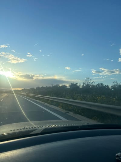 The image depicts a view from inside a vehicle, likely a car, traveling on a road. A section of road with lane markings and a roadside barrier is visible on the right side. Beyond the barrier, there's greenery, possibly trees and vegetation. The sky dominates the left side of the image, featuring a bright sun, clouds, and a distinct blue expanse. The sun's rays create glare on the windshield. The interior of the vehicle is partially visible on the right. The city of Xylofagou, Cyprus, cannot be confirmed as the location with the available information.