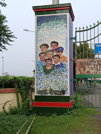 This is a medium shot, taken from a slightly low angle, of a mosaic mural on a pillar located in Benapole, Bangladesh. The mosaic depicts the faces of seven men, rendered in a pixelated style using predominantly blue, white, and flesh-toned tiles. The men are wearing various hats, including berets and what appears to be a police cap. Below the mosaic, there is text in Bengali script, also made of mosaic tiles. The pillar itself is decorated with horizontal bands of green and red tiles at its base. To the right of the pillar is a rusty green metal gate with pointed finials, partially obscuring a road and some buildings in the background. A blue sign with white text is visible behind the gate. Lush green vegetation, including vines and grass, surrounds the base of the pillar and the gate, with barbed wire visible on the left side of the frame. The sky is overcast.