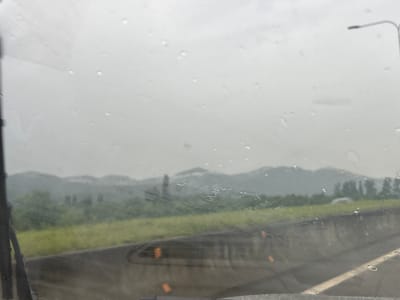 The image depicts a view through a rain-streaked windshield, looking out at a hazy landscape. In the foreground, a portion of a windshield wiper is visible on the left. The middle ground shows a grassy embankment with a concrete barrier, and a road or highway. In the distance, rolling hills or mountains are visible under an overcast sky. A street lamp is present in the upper right quadrant of the image. The scene appears to be on the outskirts of a city or in a rural area, possibly along a roadway. The weather conditions are poor due to rain. It is not possible to definitively determine the specific location within the city of ნიგოითის თემი, Georgia based solely on this image.