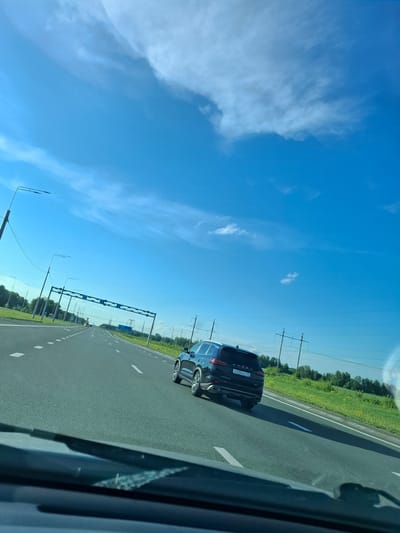 The image depicts a view from a car traveling on a highway under a clear blue sky with scattered white clouds. A dark-colored SUV is ahead on the right lane. The highway is asphalt with white lane markings. On the left side of the road, there are streetlights and a green grassy verge with trees in the distance. On the right side, there is a grassy verge with trees and utility poles. The license plate of the SUV is visible and reads "X 833 HC 55". The scene appears to be a typical highway environment, potentially in or near Ozyorki, Russia, given the license plate format and the foliage.