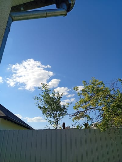 The image captures a clear, blue sky with scattered white clouds, indicative of fair weather. In the foreground, a light gray corrugated metal fence runs horizontally across the bottom of the frame. To the left, the dark shingled roof of a building is visible, with a portion of a yellow wall beneath it. A downspout extends from a building on the upper left, with a section of gutter visible. Two trees with green foliage are present; one is smaller and located towards the center-left, while a larger, more mature tree with spreading branches is positioned on the right side of the image, partially obscuring other structures in the background. The scene is likely a residential backyard in Buțeni, Moldova.